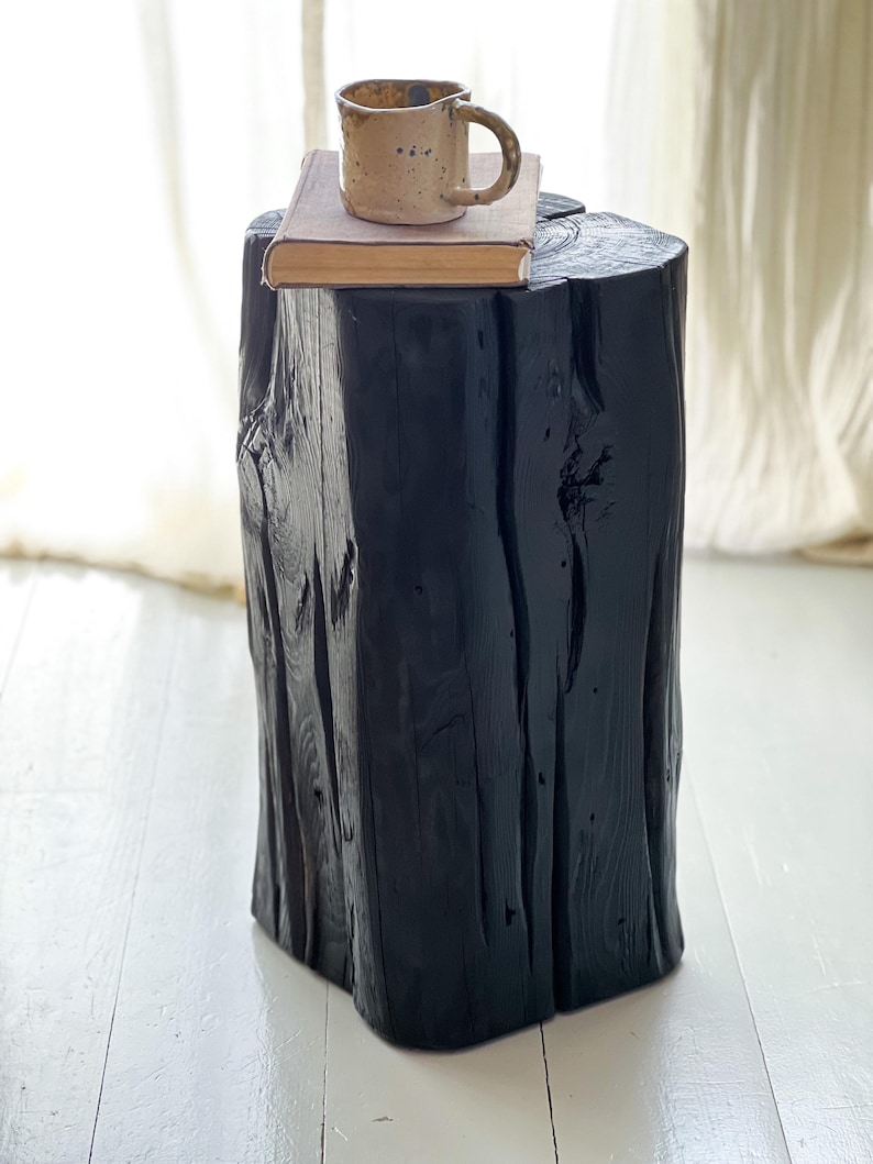 May include: A tall, cylindrical black wooden stool with a textured, natural grain finish. Resting on the flat top surface is a rectangular book with a tan cover and a speckled, light-colored ceramic mug with a handle and a dark rim.