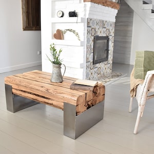 Puede incluir: Una mesa de centro de madera rústica con un acabado de madera natural y patas de metal. La mesa está hecha de madera reciclada y tiene un aspecto único y artesanal. La mesa está sobre un suelo de madera blanca en un salón.