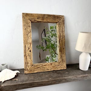 Puede incluir: Espejo rectangular con un marco de madera rústica y envejecida. El marco tiene vetas y nudos de madera visibles, lo que le da un aspecto natural y envejecido. El espejo refleja una habitación con vegetación y una lámpara.