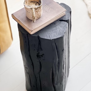 May include: A black, cylindrical wooden log stool with a textured, charred surface. Resting on top is a rectangular book with a light gray cover and a speckled, off-white ceramic mug filled with a dark liquid, featuring a prominent handle.