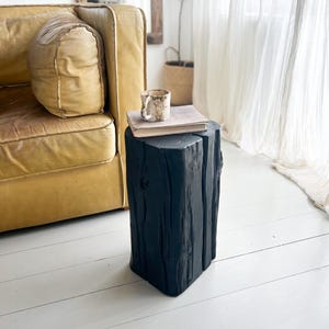 May include: A black wooden stool with a vertical grain texture and a flat top surface. A beige ceramic mug and a closed book with a light-colored cover are placed on top of the stool, which stands on a white floor next to a yellow leather sofa.