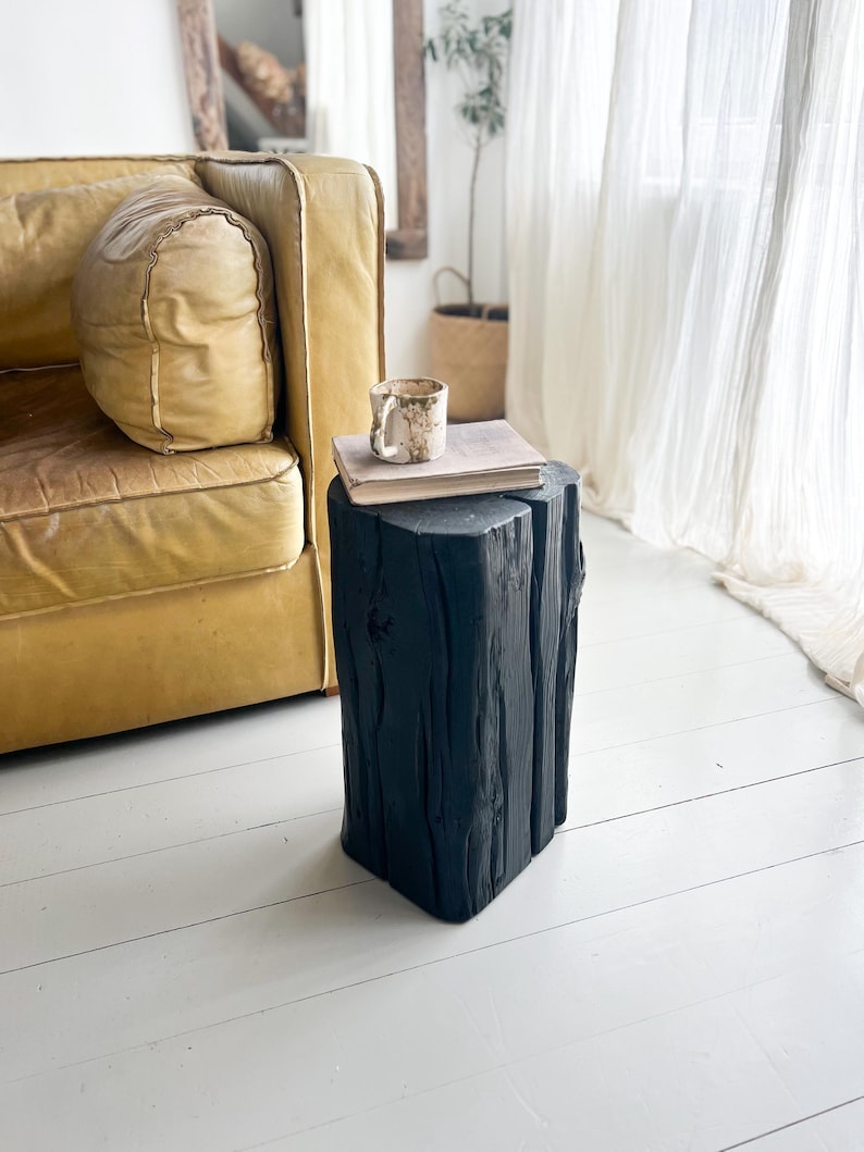 May include: A black wooden stool with a vertical grain texture and a flat top surface. A beige ceramic mug and a closed book with a light-colored cover are placed on top of the stool, which stands on a white floor next to a yellow leather sofa.