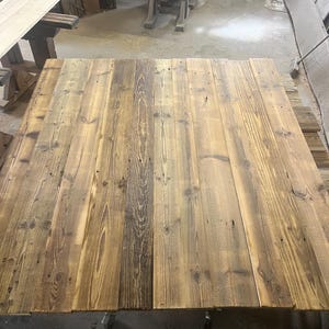 May include: A rectangular tabletop constructed from multiple joined wooden planks. The wood features a natural grain pattern with varying shades of brown, ranging from light tan to dark espresso, and includes visible knots and wood texture.