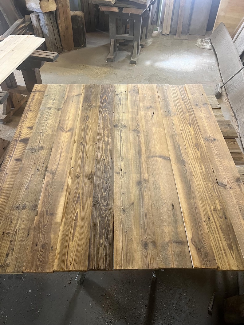 May include: A rectangular tabletop constructed from multiple joined wooden planks. The wood features a natural grain pattern with varying shades of brown, ranging from light tan to dark espresso, and includes visible knots and wood texture.