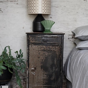Puede incluir: Una mesita de noche de estilo industrial con acabado metálico envejecido. Una pantalla de lámpara estampada se asienta sobre una base de lámpara negra. Un jarrón de cristal verde añade un toque de color. También se ven una planta en maceta y una cama con ropa de cama a rayas.