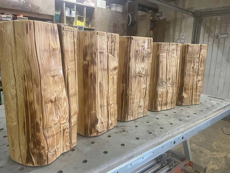 May include: A row of six vertical wooden logs with a natural light brown finish, featuring visible wood grain, knots, and vertical cracks. Each log has a rectangular shape with a flat top and a small rectangular brand tag attached to the upper edge.