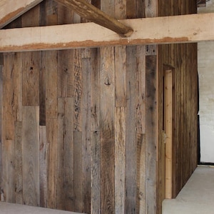 May include: A wall constructed from vertical reclaimed wood planks featuring varied brown and gray tones with visible wood grain and knots. The structure includes a doorway and is framed by large, light-colored horizontal wooden beams.
