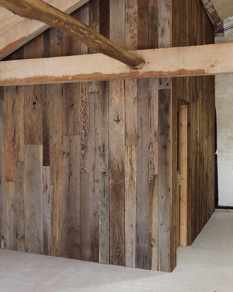 May include: A wall constructed from vertical reclaimed wood planks featuring varied brown and gray tones with visible wood grain and knots. The structure includes a doorway and is framed by large, light-colored horizontal wooden beams.