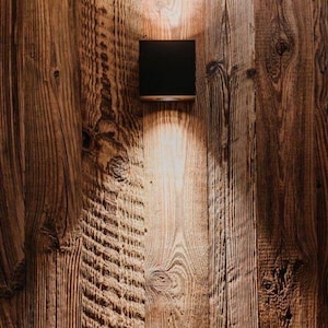 May include: A square black wall-mounted light fixture is attached to a vertical wooden plank wall. The lamp emits a focused beam of warm light that illuminates the textured grain of the wood planks directly below the fixture.