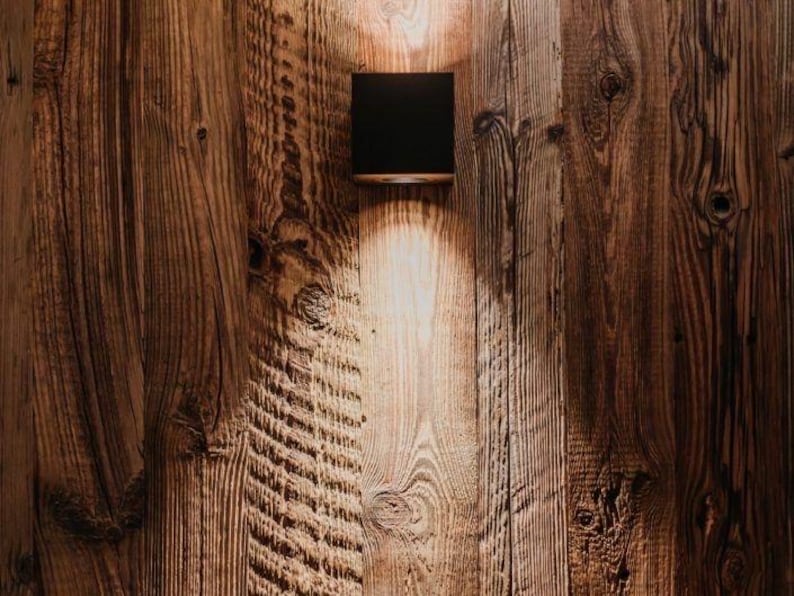 May include: A square black wall-mounted light fixture is attached to a vertical wooden plank wall. The lamp emits a focused beam of warm light that illuminates the textured grain of the wood planks directly below the fixture.