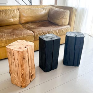 May include: Three vertical wooden stools with a square cross-section and a central vertical split. One stool features a natural light brown wood grain finish, while the other two stools are finished in a solid matte black color.