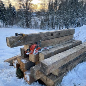 May include: A stack of weathered, rectangular brown timber beams arranged in a pile. An orange and black chainsaw with a silver metal guide bar rests on the center beam. A small, yellow and black cordless drill sits on the top beam.