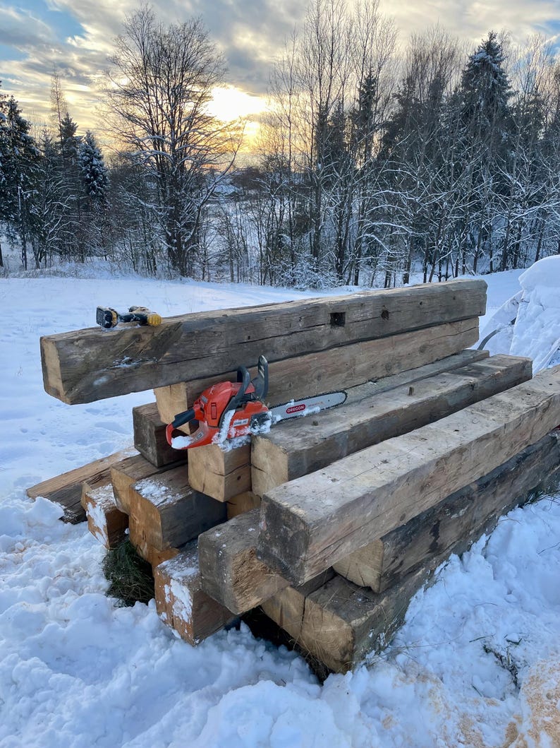 May include: A stack of weathered, rectangular brown timber beams arranged in a pile. An orange and black chainsaw with a silver metal guide bar rests on the center beam. A small, yellow and black cordless drill sits on the top beam.