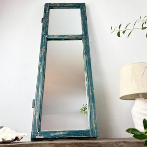 May include: A tall, rectangular mirror with a distressed teal frame. The mirror is divided into two sections, with a smaller top section and a larger bottom section. The frame has a weathered, rustic appearance, with visible wood grain and chipped paint.