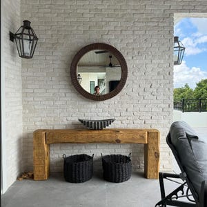 Puede incluir: Una consola rústica de madera con dos cestas negras tejidas debajo de un espejo redondo con marco tejido. La pared es de ladrillo blanco y hay dos faroles negros. Un cuenco decorativo de metal está sobre la mesa.