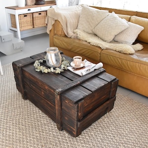 Può includere: Un tavolino da caffè in legno marrone scuro con design a baule e angoli in metallo. È posizionato su un tappeto beige. Una tazza di caffè, una candela in un barattolo di vetro e una ghirlanda decorativa sono sul tavolo. Il tavolo misura circa 100 cm di lunghezza.