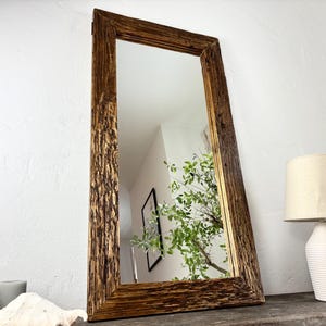 Peut inclure: Un miroir rectangulaire avec un cadre en bois rustique et texturé. Le cadre a un aspect naturel et vieilli avec des nuances de brun. Le miroir reflète une pièce avec de la verdure et une lampe.