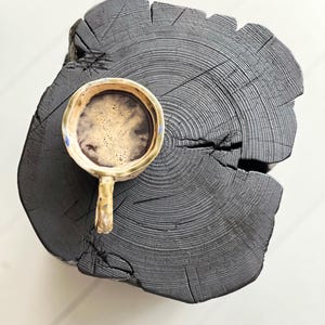 May include: A ceramic mug with a speckled beige and blue glaze filled with dark coffee, resting on a large, circular, charred black wooden log slice that displays prominent natural wood grain rings and a deep structural crack.