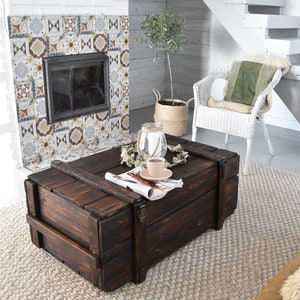 May include: A dark brown wooden coffee table with metal clasps and a rectangular shape. The table, placed on a light rug, has a cup of coffee and a newspaper on top. The table has a rustic, vintage aesthetic, and is approximately 120 cm long.