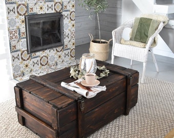 May include: A dark brown wooden coffee table with a rectangular shape and metal hardware. The table is placed on a light-colored rug, with a cup of coffee and a newspaper on top. The table has a rustic, vintage aesthetic.