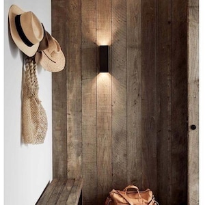 May include: A rectangular black wall sconce mounted on a vertical wood plank wall, with a wooden bench, a tan leather duffel bag, and two straw hats with a woven mesh bag hanging on a white wall nearby in an entryway space.