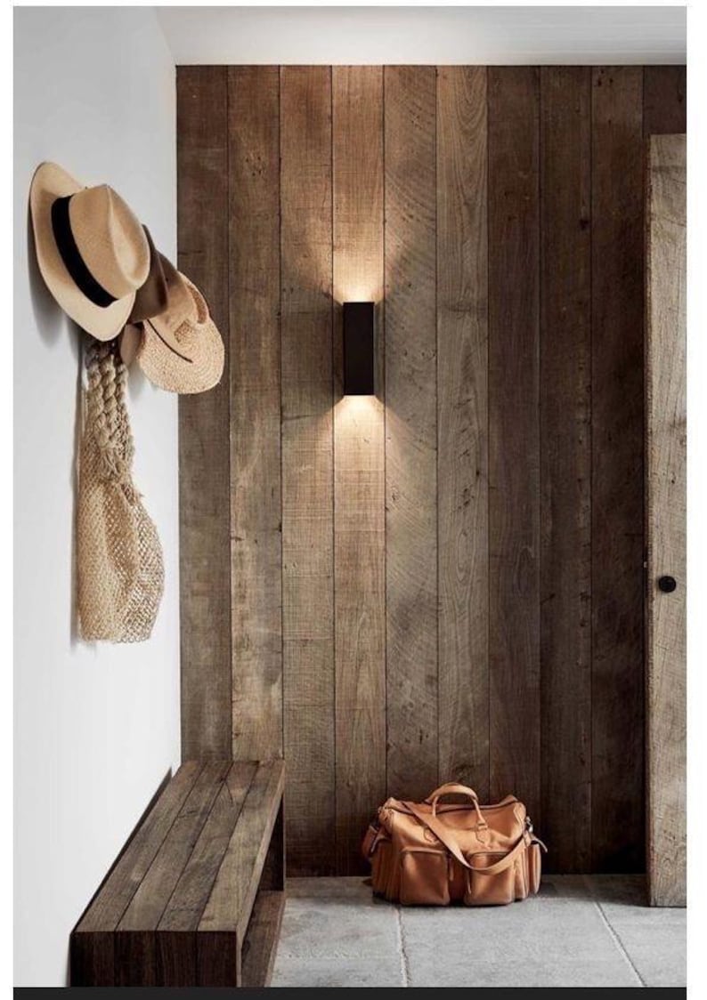 May include: A rectangular black wall sconce mounted on a vertical wood plank wall, with a wooden bench, a tan leather duffel bag, and two straw hats with a woven mesh bag hanging on a white wall nearby in an entryway space.