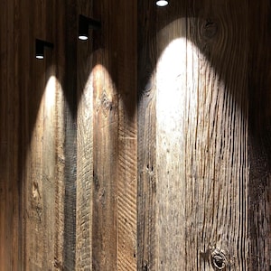 May include: Three small black cylindrical wall-mounted spotlights are attached to a vertical wooden wall paneling. Each light fixture casts a bright, focused beam of white light downward onto the textured, weathered brown wood planks.