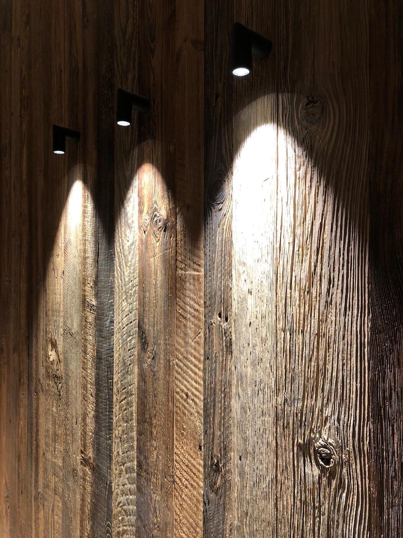 May include: Three small black cylindrical wall-mounted spotlights are attached to a vertical wooden wall paneling. Each light fixture casts a bright, focused beam of white light downward onto the textured, weathered brown wood planks.