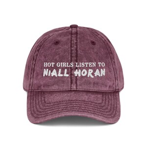May include: A maroon baseball cap with a curved brim. The cap features the white text "HOT GIRLS LISTEN TO NIALL HORAN" embroidered on the front. The hat has a vintage, worn look.