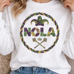Könnte beinhalten: Weißes Sweatshirt mit einem kreisförmigen Design. Das Design zeigt das Wort "NOLA" in Lila, Gold und Grün, eine Narrenkappe, gekreuzte Schlüssel und eine Krone, alles innerhalb eines geflochtenen Seilrandes. Ein lässiger Sweatshirt-Stil.