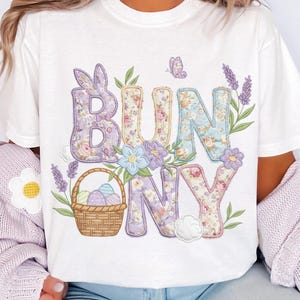 Könnte beinhalten: Weißes T-Shirt mit dem Wort "BUNNY" in Buchstaben mit Blumendruck. Das Design umfasst einen Korb mit Ostereiern, Lavendelzweigen, einen Schmetterling und dekorative Blumen. Die Buchstaben haben ein Patchwork-Design.