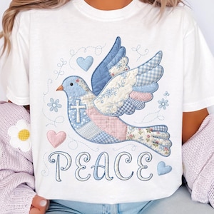 Könnte beinhalten: Weißes T-Shirt mit einem Patchwork-Tauben-Design in Blau, Rosa und Weiß. Die Taube hat ein Kreuz und ist von Herzen und Blumen umgeben. Das Wort "PEACE" ist darunter in Schrift geschrieben.