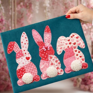 Könnte beinhalten: Türkisfarbener Stoff mit drei applizierten Osterhasen. Jeder Hase ist ein Patchwork aus roten und rosa Stoffen mit Herz- und XOXO-Mustern. Weiße Pompon-Schwänze und eine rosa Schleife vervollständigen das Design.