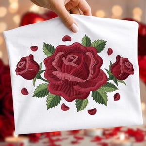 Könnte beinhalten: Weißer Stoff mit einem gestickten Design einer großen roten Rose, zwei kleineren Rosenknospen, grünen Blättern und verstreuten roten Blütenblättern. Der Hintergrund ist verschwommen.
