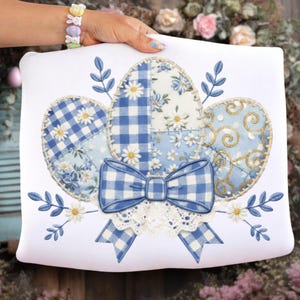 Könnte beinhalten: Weißes Kissen zum Thema Ostern mit drei dekorativen Eiern in Blautönen. Die Eier haben Patchwork-Designs mit Blumen- und Vichy-Mustern. Eine blaue Vichy-Schleife und eine Spitzentrimmung befinden sich unten, mit gestickten Blättern und Blumen.