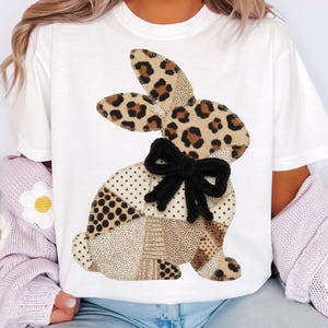 Könnte beinhalten: Weißes T-Shirt mit einem Patchwork-Hasen-Design. Der Hase hat Leopardenohren, eine schwarze Schleife und einen Körper aus verschiedenen gemusterten Stoffen in Braun- und Beigetönen. Ein lässiger Artikel für den Alltag.