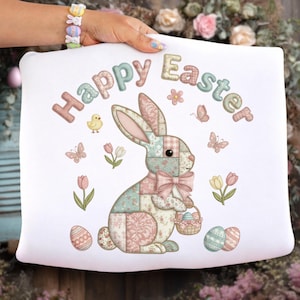 Könnte beinhalten: Weißes Kissen mit einem pastellfarbenen Patchwork-Hasen-Design, den Worten "Happy Easter" und Ostermotiven. Der Hase hält einen Korb mit Eiern, umgeben von Schmetterlingen, Blumen und verzierten Eiern. Das Kissen wird von einer Person gehalten.