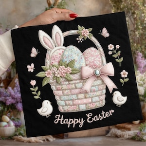 Könnte beinhalten: Schwarzes quadratisches Kissen mit einem Osternest-Design. Der Korb enthält Ostereier, Hasenohren und eine rosa Schleife. Die Worte "Happy Easter" sind in weißer Schrift geschrieben. Schmetterlinge und Blumen sind ebenfalls abgebildet.