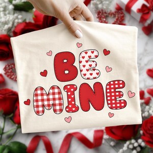 Könnte beinhalten: Ein cremefarbenes T-Shirt mit den Worten "BE MINE" in Rot, mit Herzmuster und Karomuster. Kleine rosa Herzen sind um den Text herum verstreut. Das T-Shirt wird von einer Hand gehalten, mit Rosen und Bändern im Hintergrund.