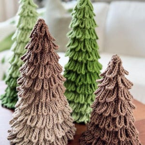 May include: Four knitted Christmas tree decorations in shades of green and brown. The trees have a textured, layered design, with a cone shape. The trees are arranged on a wooden surface, with a blurred background.