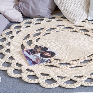 May include: A cream-colored crocheted round rug with a delicate lace pattern. The rug is on a gray surface with pillows and a magazine on top.
