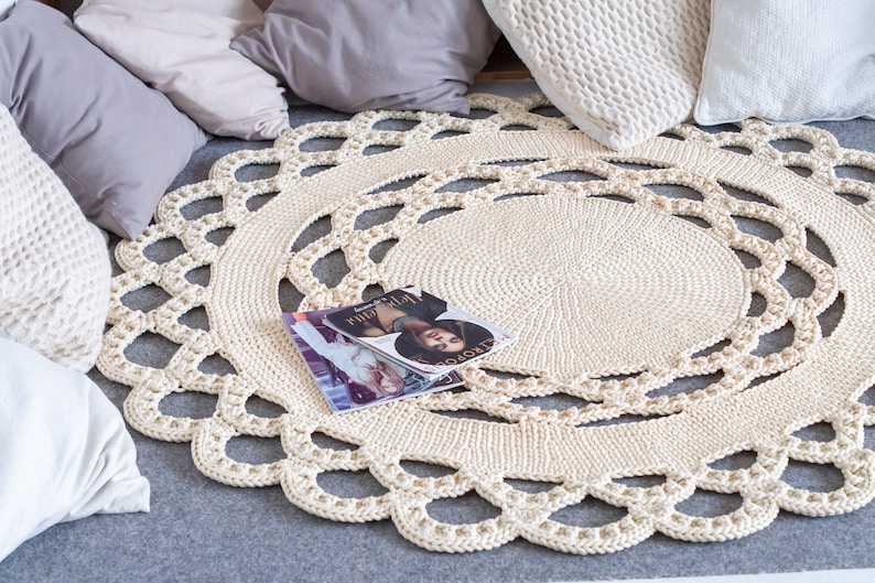 May include: A cream-colored crocheted round rug with a delicate lace pattern. The rug is on a gray surface with pillows and a magazine on top.