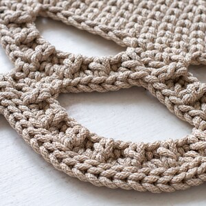 May include: Close-up of a beige crocheted rug with a unique openwork design. The rug is made of thick, textured yarn and has a natural, earthy feel.