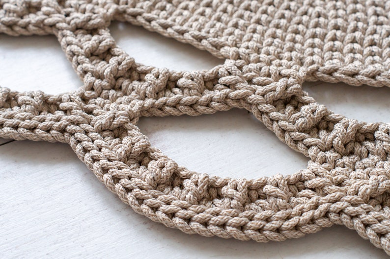 May include: Close-up of a beige crocheted rug with a unique openwork design. The rug is made of thick, textured yarn and has a natural, earthy feel.