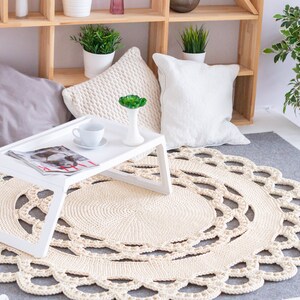 May include: A white crocheted round rug with a decorative border sits on a gray rug in a living room setting. A white tray table with a cup and saucer and a magazine sits on the rug.  There are white pillows and a wooden shelf in the background.