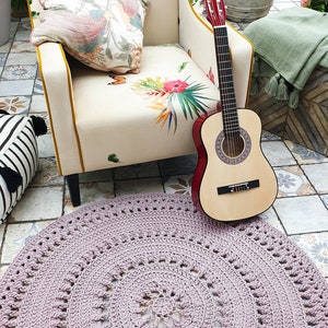 Puede incluir: Una alfombra redonda de crochet rosa claro con un patrón circular. La alfombra está delante de un sillón con estampado floral y una guitarra acústica roja.