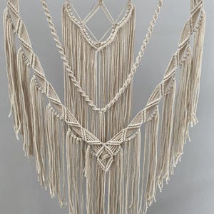 以下が含まれることがあります： ベージュのマクラメの壁掛け。複雑な結び目の模様と長いフリンジが特徴です。このテキスタイルアートは、天然の木の枝から吊り下げられており、ボヘミアンスタイルを表現しています。デザインは幾何学的な形と滝のように流れる糸が特徴です。