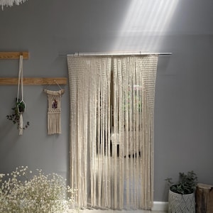 May include: A cream-coloured macrame curtain hangs in front of a window. The curtain is made of knotted cords, creating a textured, bohemian look. The room has grey walls and a plant in a macrame pot.