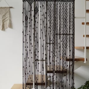Könnte beinhalten: Schwarzer Makramee-Vorhang mit Perlenakzenten, der vor einer Treppe hängt. Ein cremefarbenes Makramee-Wandbehang ist links an der Wand angebracht. Der Vorhang besteht aus geknoteten Kordeln.