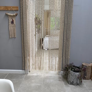 May include: Beige macrame curtains hanging in a doorway. The curtains are made of knotted rope, with a fringe at the bottom. A macrame wall hanging is on the left. A potted plant sits to the right.